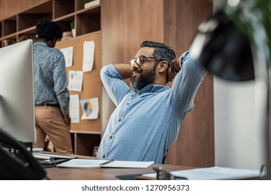 Happy Middle Eastern Businessman Relaxing In His Office While Looking Computer Monitor. Smiling Mature Business Man With Beard And Eyeglasses Sitting With Hands Behind Head At Office Desk.