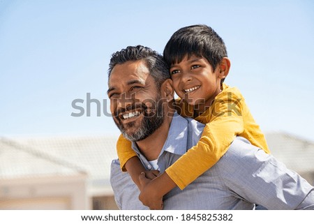 Happy middle eastern child enjoying ride on father back outdoor. Smiling dad giving piggyback ride to son on street while looking away with copy space. Indian cheerful man carrying on shoulder kid.
