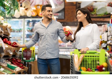 Happy Middle Eastern Couple Buying Food Choosing Fresh Vegetables During Grocery Shopping In Modern Supermarket. Spouses Purchasing Groceries In Local Market. Consumerism