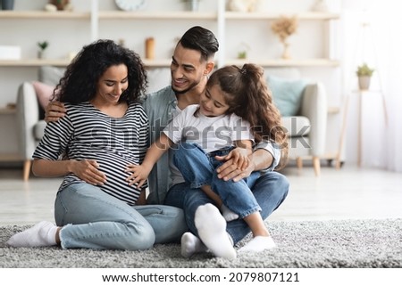 Happy middle eastern family father, pregnant mother and preschool kid posing together, sitting on floor at home, waiting for baby, cute little girl touching her expecting mom big tummy, copy space