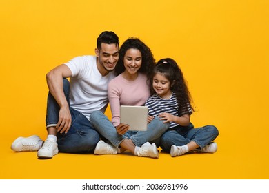 Happy Middle Eastern Family Of Three With Digital Tablet Relaxing On Floor On Yellow Studio Background, Cheerful Arab Parents And Little Daughter Shopping Online Or Watching Movie Together