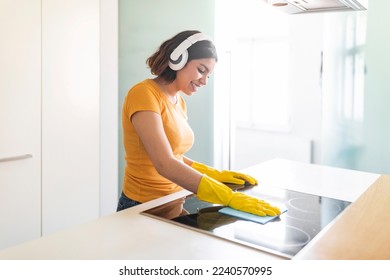 Happy Middle Eastern Female Listening Music In Headphones While Cleaning Kitchen, Cheerful Young Arab Woman Polishing Induction Cooktop Surface With Rag And Enjoying Favorite Songs, Copy Space