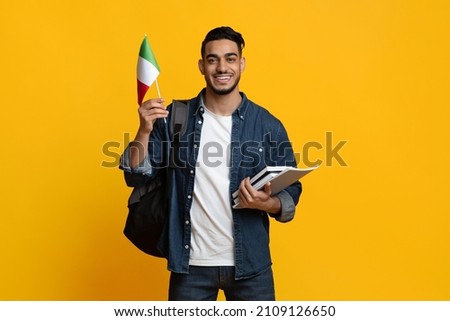 Happy middle eastern guy student with backpack and bunch of books showing flag of Italy over yellow studio background, copy space. Positive young man studying abroad at Italian university