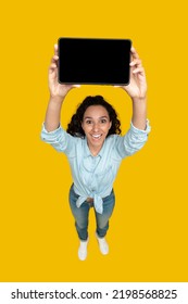 Happy Middle Eastern Lady Showing Digital Tablet With Empty Screen Advertising Website Posing Standing Over Yellow Studio Background. Vertical Shot, High Angle Shot