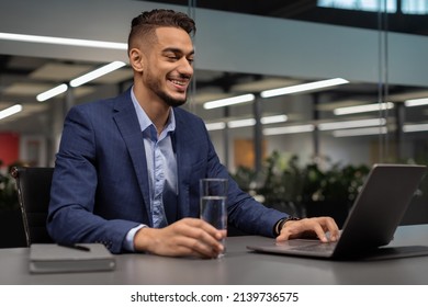 Happy middle eastern man in suit manager drinking water while working on laptop, typing on computer keyboard, making marketing research or sending emails, office interior, copy space - Powered by Shutterstock