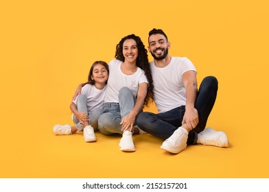 Happy Middle Eastern Parents And Little Daughter Embracing Smiling To Camera Sitting Together On Yellow Background In Studio. Shot Of Cheerful Arabic Family. Full Length