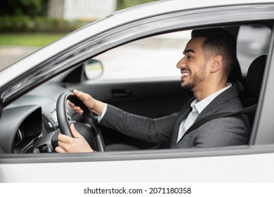 Happy middle eastern young businessman driving new auto, going to office, shot from outside, side view. Handsome successful arab guy manager going home after good day, looking at road and smiling