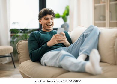 Happy Middle Eastern Young Man Using Cellphone And Laughing Texting Lying On Sofa At Home. Cheerful Man Holding Smartphone Playing Game And Networking Online Having Fun On Weekend - Powered by Shutterstock