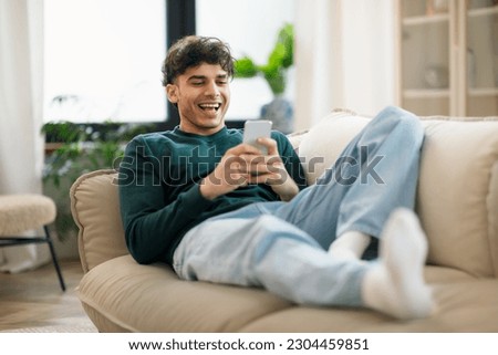 Happy Middle Eastern Young Man Using Cellphone And Laughing Texting Lying On Sofa At Home. Cheerful Man Holding Smartphone Playing Game And Networking Online Having Fun On Weekend