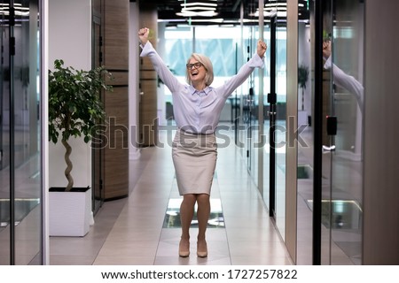 Happy middle-aged business lady celebrates personal business goal achievements standing dancing in corridor office hallway raised hands scream with joy feels excited. Growth progress in career concept