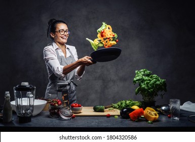 Happy Middle-aged Female Chef Tossing Chopped Vegetables In The Air From A Frying Pan. Healthy Food Concept. Studio Photo On A Dark Background