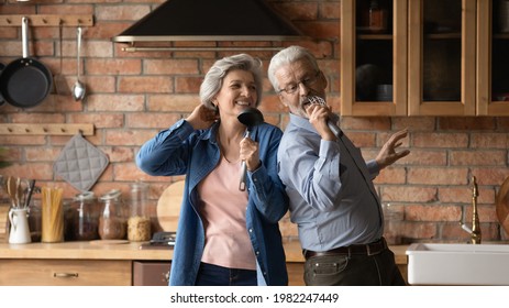 Happy middle-aged hoary wife and husband dancing in kitchen listen music singing song using kitchenware like microphones enjoy karaoke together feel carefree. Hobby, untroubled retired life concept
