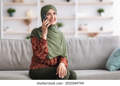 Happy Middle-Aged Islamic Woman In Hijab Talking On Cellphone Sitting On Sofa In Living Room Indoor. Muslim Lady Chatting Communicating Via Smartphone At Home. Pleasant Phone Call, Communication