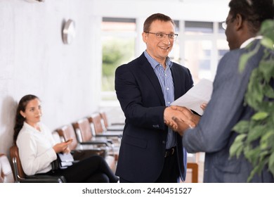 Happy middle-aged man holding a document shaking hands with a person in waiting hall. Visitor sitting on chair and watching them - Powered by Shutterstock