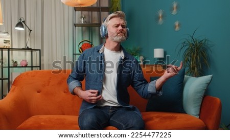 Happy middle-aged mature rocker man in wireless headphones relaxing at home dancing on couch listening energetic disco dancing music, playing on imaginary guitar. People weekend leisure activities