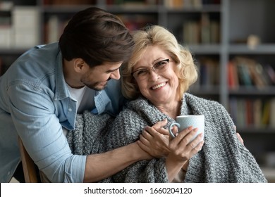 Happy middle-aged mother relax in chair drink tea enjoy family weekend reunion with grown-up son, smiling senior 70s mom rest at home spend time with caring adult man child, bonding concept
