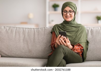 Happy Middle-Aged Muslim Woman Using Smartphone Texting Reading Messages And Browsing Internet Sitting On Couch At Home, Wearing Glasses And Hijab. People and Gadgets Concept