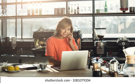 Happy Middle-aged Woman Barista Working On Laptop And Talking On Smartphone, Accepting An Order At Modern Coffee Shop. Small Business, Online Purchase And Service Concept, Copy Space