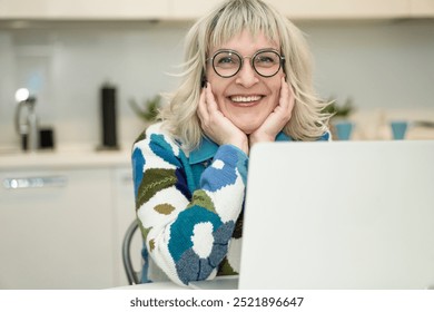 Happy middle-aged woman in glasses smiling cheerfully looking at camera working on laptop computer against background of modern kitchen with copy space - Powered by Shutterstock