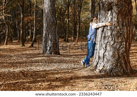 Happy middle-aged woman hugging big tree in autumn forest. Spending time in fall nature for exercise and fitness for wellness and healthy lifestyle. Freedom and unity with nature concept.