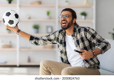 Happy Middle-eastern Man Football Fan Watching Game On TV At Home, Emotional Arab Guy Sitting On Couch With Soccer Ball And Remote, Raising Hand Up And Screaming, Celebrating His Team Win