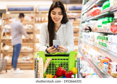 Happy Middle-Eastern Woman Using Smartphone In Supermarket Doing Grocery Shopping Walking With Shop Cart Indoors. Groceries Shopping Mobile App For Your Phone. Selective Focus