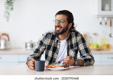 Happy middle-eastern young bearded man in casual outfit and stylish glasses enjoying healthy lunch at home, sitting at kitchen table, eating breakfast, looking at copy space, healthy deit concept - Powered by Shutterstock