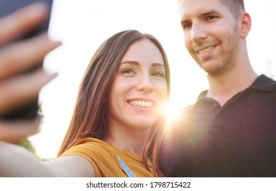 Happy Millenial Couple Taking Selfie Outdoor In Sunset - Smiling Friends Using Smartphone In Backlight 