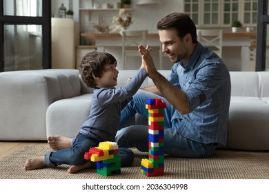 Happy millennial 30s dad giving praise and high five to little son for building toy tower from construction plastic blocks. Joyful kid playing learning games with daddy on heating floor
