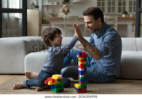 Happy\
millennial 30s dad giving praise and high five to little son for\
building toy tower from construction plastic blocks. Joyful kid\
playing learning games with daddy on heating\
floor