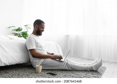 Happy millennial african american bearded guy sit near bed work on laptop in bedroom interior on window background, side view. Chat in social networks, business at home, new normal and social distance