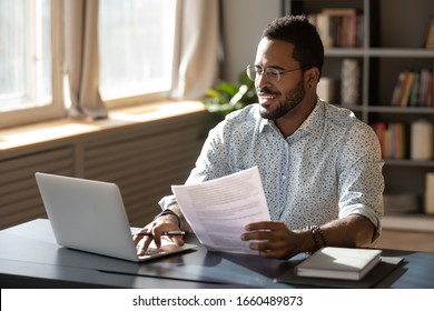 Happy millennial african american businessman in eyewear holding documents, doing paperwork, preparing report or analyzing market research results, working on computer in modern workplace office.