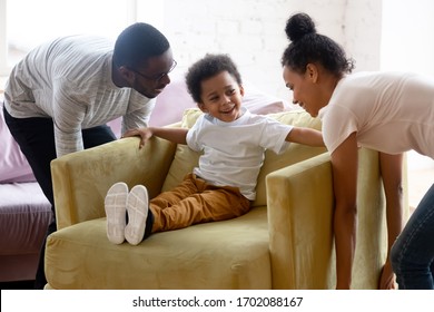 Happy millennial african american couple moving armchair with little son. Diverse smiling family of woman, man and sitting boy playing together carry chair and have fun.