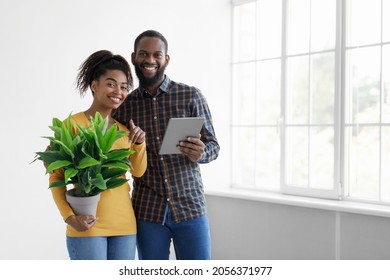 Happy Millennial African American Couple With Potted Plant Hold Tablet And Looking At Camera Near Window In Room Interior. Digital Device, App For Flat Design, Moving And Home Renovation, Free Space