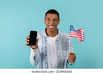 Happy Millennial African American Guy Show Smartphone With Blank Screen And USA Flag, Isolated On Blue Background, Studio. New App For Study, Learning English, Communication And Chatting Online Remote