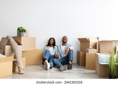 Happy millennial african american spouses relocating to new apartment, sitting among cardboard boxes at new home and smiling with closed eyes, empty space. Mortgage and ownership