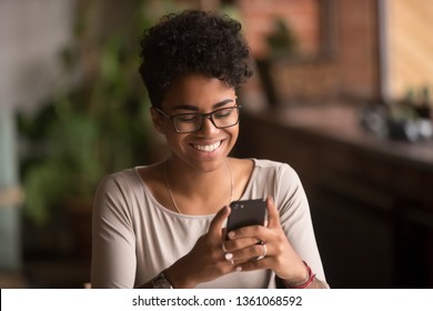 Happy millennial african american woman holding smartphone using ecommerce apps, playing mobile games, chatting in messengers or social media networks, surfing internet or texting message on phone