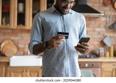 Happy millennial African guy shopping with mobile phone, ordering goods on internet, paying for purchase by credit card, using online banking app for transferring money, getting good cash back