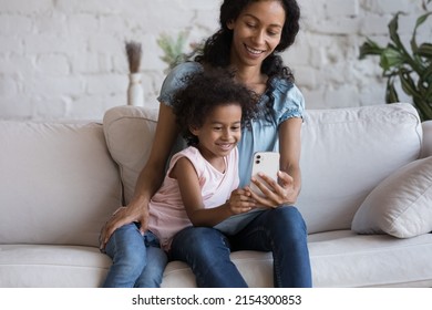 Happy Millennial African Mom And Cheerful Gen Z Daughter Kid Making Video Call On Smartphone, Taking Selfie Picture, Shopping Online On Mobile Phone, Using Learning App On Cell, Relaxing At Home