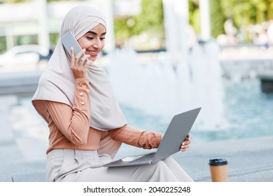 Happy millennial arab islamic female in hijab digital nomad calls on smartphone, work remote on laptop outdoors. Business, lifestyle, remote job in city, during social distancing at covid-19 pandemic
