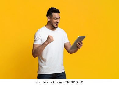 Happy millennial arabic guy with digital tablet celebrating success, looking at pad screen and raising fist up, lucky young man gambling online, using newest mobile app, yellow background, copy space