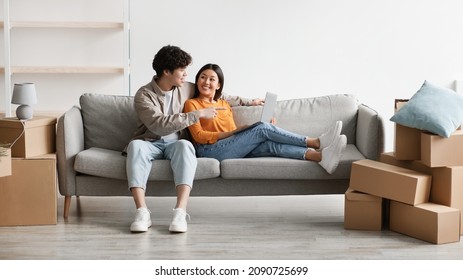 Happy Millennial Asian Couple With Laptop Sitting On Couch Among Cardboard Boxes And Planning Design Of Their New Home. Young Family Browsing Internet For House Decoration Ideas, Banner