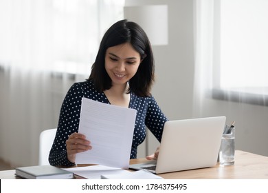 2000年の幸せなアジア人女性がノートパソコンのデスクに座り込み、紙の通信や手紙の書類で良い知らせを読むことを考え、にこやかなベトナム人の若い女性がコンピューターの準備に関する報告書を調べる