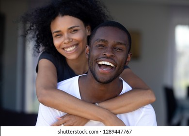 Happy Millennial Black Couple Laughing Having Fun Outdoors, Excited African Man Piggybacking Woman Standing Outside New House On Terrace Looking At Camera, Young American Family Headshot Portrait