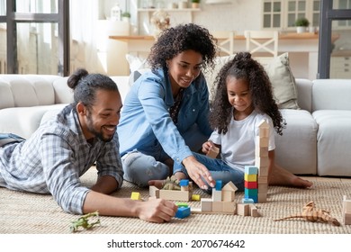 Happy millennial Black couple of parents and cute daughter kid playing on heating carpeted floor at cozy home, constructing town, buildings, towers from toy wooden bricks. Family game activity