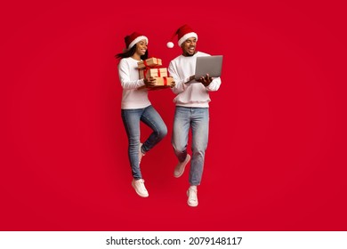 Happy millennial black couple in red Santa hats buying xmas gifts for relatives and friends on Internet via notebook, jumping up and smiling over red studio background, enjoying super offers - Powered by Shutterstock