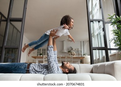 Happy Millennial Black Father And Excited Daughter Kid Playing Airplane On Couch. Daddy Holding Lifting Girl With Plane Wing Hands Up In Air. Carefree Child Looking Forward. Family Leisure