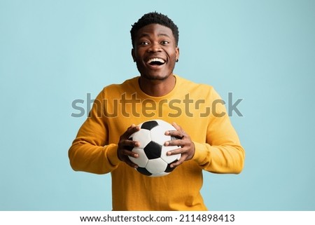 Happy millennial black guy in casual with soccer ball posing on blue studio background, emotional african american young man football fan going to game, copy space. Sport concept