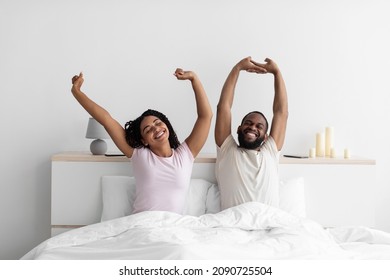 Happy millennial black husband and wife woke up, stretching bodies on white bed in minimalist bedroom interior. Couple in white t-shirts in morning after wake up. Healthy sleep, vacation and weekend