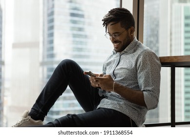 Happy Millennial Black Man Sit By Big Window At Modern Studio Apartment Having Virtual Dating Flirting With Beloved Female By Phone. Smiling African American Guy Spend Leisure Time At Home Using Cell